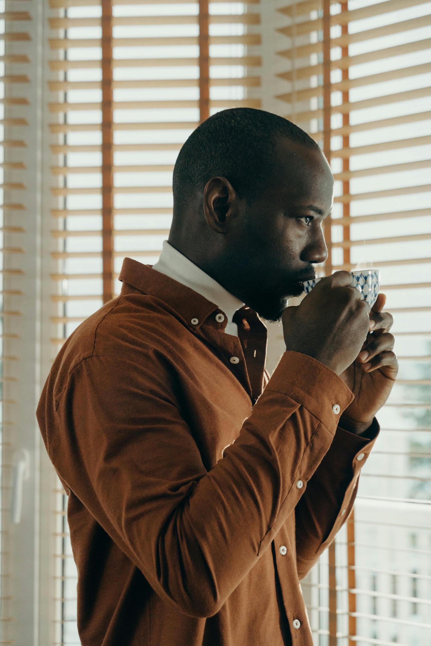 Man in a brown jacket drinking from a cup in front of window blinds